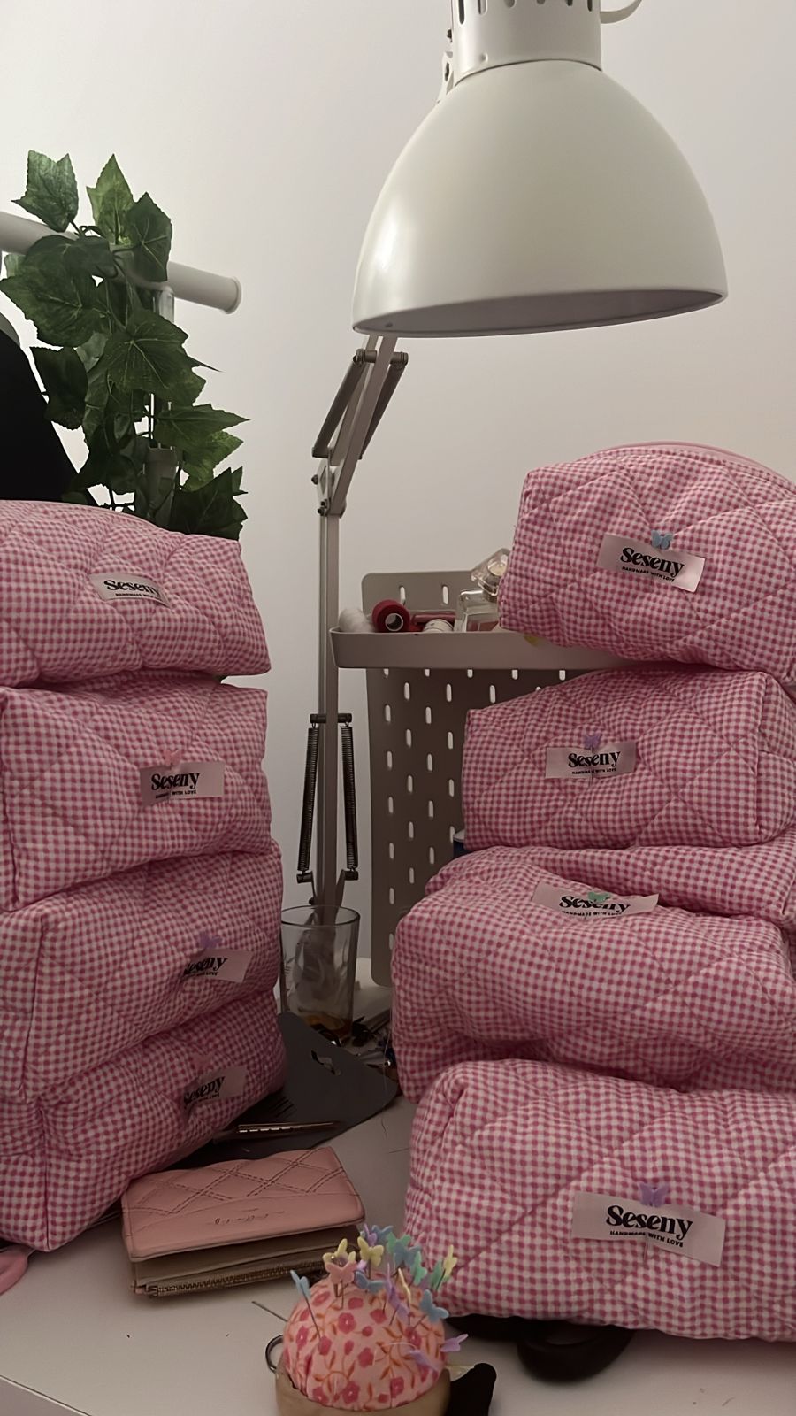 Trousse Seseny en tissu vichy rose, empilées sur une table avec lampe et plante d'intérieur.